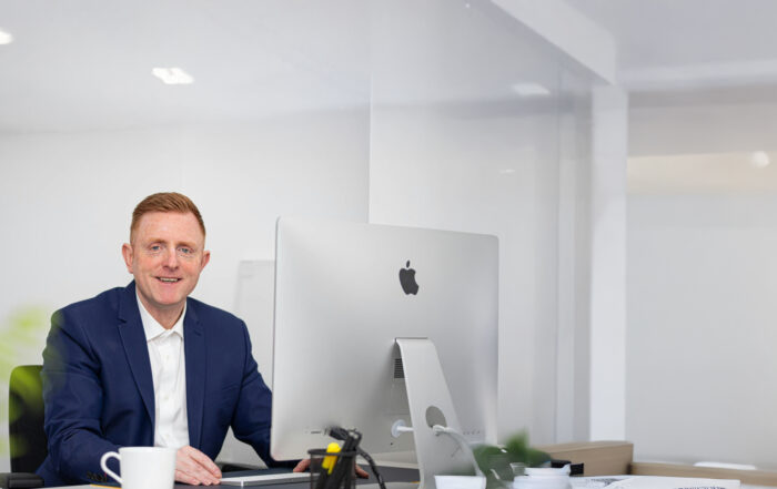 Ein Geschäftsführer im blauen Anzug sitzt an seinem Schreibtisch vor einem iMac und lächelt in die Kamera – Symbol für moderne CEO-Kommunikation auf LinkedIn.